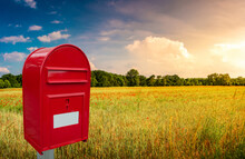 Big Red Cozy Postbox With White Empty Note Space For Address Is Standing Outdoor In Front Of Beautiful Countryside Landscape At Sunset Background With Farm Field And Poppies Flowers.