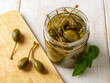 Marinated caper fruits in an open glass jar on a wooden table. Picled caper berries for condiment and garnish. Canned caperberry. Mediterranean cuisine ingredient. Tasty pickles.