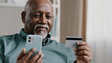 Elderly African Dominican old mature man at home online transaction buying hold smartphone credit card successful payment buy on internet with mobile app food delivery order with phone easy e-payment