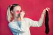 a beautiful girl in pink glasses on a pink background with surprise and disgust holds a dirty black men's sock in her hands and covers her nose from the smell