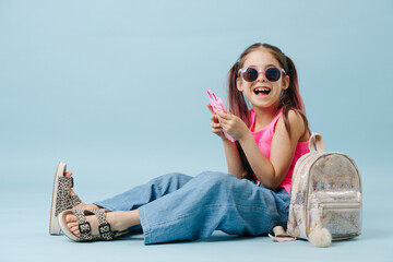 Cheerful tween girl in pink shirt and round sunglasses using her phone. Sitting on the floor over blue background.