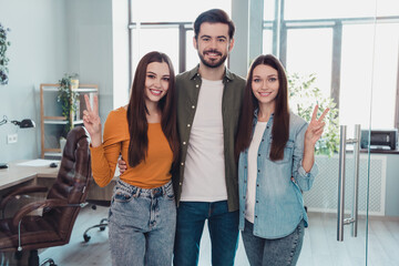 Poster - Portrait of three positive cheerful people employee hug hand fingers demonstrate v-sign office indoors