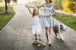 European girl mother hipster with tattoo holds hands, helps teach child daughter to ride skateboard. dog Australian Shepherd breed sidewalk in park, warm summer evening and family time and hobbies
