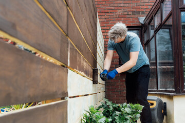 Young man worker paints with a roller a wooden board fence in the garden. DIY, Do it yourself concept. House improvement. Home renovation and refurbishment. Selective focus, copy space.