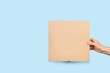 Mano de hombre sosteniendo una caja de cartón marrón ecológica de pizza sobre un fondo celeste liso y aislado. Vista de frente y de cerca. Copy space