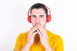 © Roquillo - Shocked Young caucasian man wearing yellow t-shirt over white background stares fearful at camera keeps mouth widely opened wears wireless stereo headphones on ears