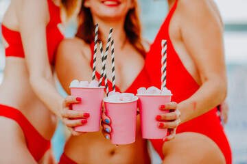 Three pretty women in red swim suits holds pink paper cups with straws, making cheers at the beach. Focus is at cups