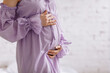 Side view close up cropped of pregnant woman in purple dress touching belly tummy, expecting baby, young future mother standing at home on white brick background, copy space, motherhood concept