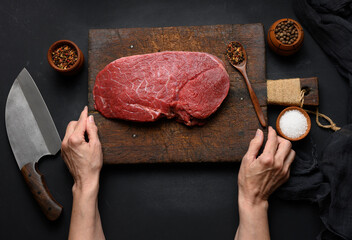 Raw beef tenderloin lies on a wooden cutting board and spices for cooking on a black table, top view and large kitchen knife