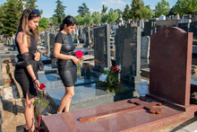 Mother And Her Daughter In Grief, In Black Clothes, Holding A Flower And Mourning A Deceased Loved One On Cemetery