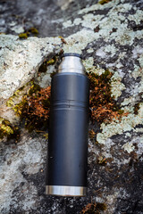 Wall Mural - Metal black thermos close-up lies on a stone, vacuum bottle for hot tea, thermos without lid, gray moss yagel, camping equipment.