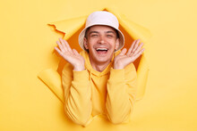 Portrait Of Happy Excited Amazed Man Wearing Casual Hoodie And Panama Looking Through Torn Hole In Yellow Paper, Looking At Camera With Happy Smile, Raised Arms, Expressing Positive Emotions.