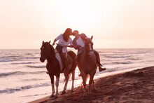 A Loving Couple In Summer Clothes Riding A Horse On A Sandy Beach At Sunset. Sea And Sunset In The Background. Selective Focus 