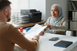 © AnnaStills - Modern Muslim woman wearing hijab working in embassy sitting at desk in front of Caucasian man giving him documents after visa approval