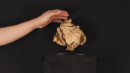 The hand and a crumpled brawn paper maul and trash can on black background.