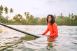 Black long-haired teen man floating on long surfboard, waiting for a wave ready for surfing with palm grove litted sunset rays. Extreme water sports or traveling to exotic countries concept. Sri Lanka