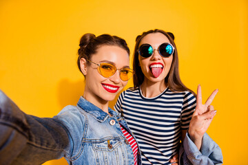 Wall Mural - Portrait of two positive carefree girls take selfie show tongue v-sign isolated on yellow color background