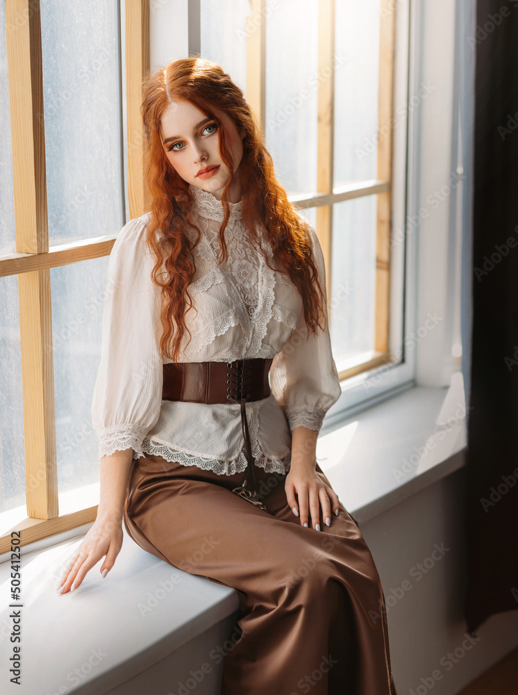 Red-haired woman in vintage dress sits on large classic window waiting  love. Clothing costume countess old art style white blouse, brown long  skirt. Curly red hair. Redhead girl princess 1800s stylish Stock-foto |