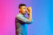 Side view portrait of handsome young adult man in shirt looking far away through monocular gesture, spying shocking content. Indoor studio shot isolated on colorful neon light background.