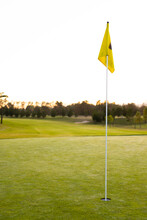 Yellow Golf Flag In Hole Amidst Grassy Landscape Against Trees And Clear Sky During Sunset