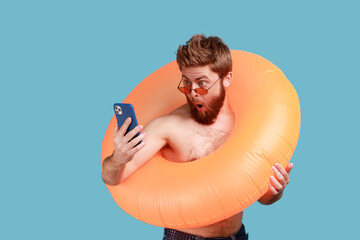 Portrait of astonished surprised bearded man in sunglasses standing with orange rubber ring and holding cell phone, looking at display with open mouth. Indoor studio shot isolated on blue background.