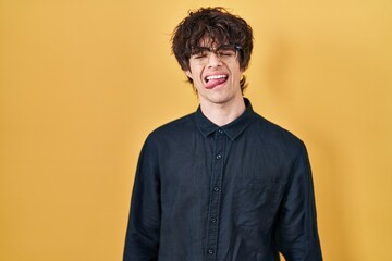 Poster - Young man wearing glasses over yellow background sticking tongue out happy with funny expression. emotion concept.