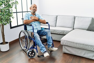 Poster - Handsome senior man sitting on wheelchair at the living room smiling with hands on chest with closed eyes and grateful gesture on face. health concept.