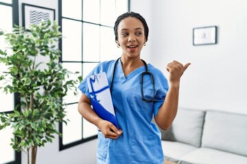 Poster - Young african american woman working wearing doctor uniform working at the clinic pointing thumb up to the side smiling happy with open mouth