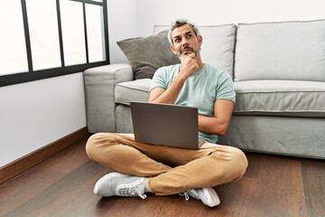 Poster - Middle age hispanic man using laptop sitting on the floor at the living room with hand on chin thinking about question, pensive expression. smiling with thoughtful face. doubt concept.