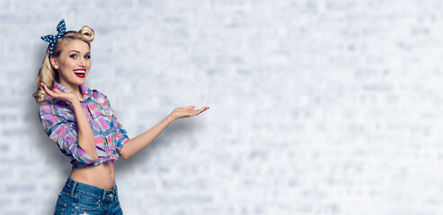 Wall Mural - Very happy woman holding or giving. Excited girl in pin up cloth, showing some product or copy space for advertise slogan text. Retro fashion and vintage. White brick wall background. Wide composition