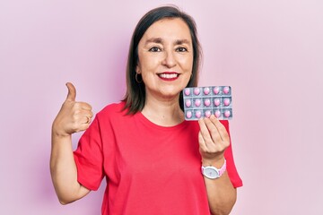 Poster - Middle age hispanic woman holding pills smiling happy and positive, thumb up doing excellent and approval sign