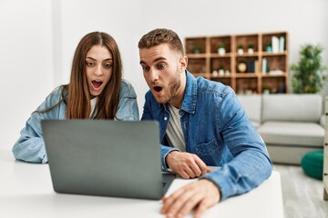 Wall Mural - Young caucasian couple working using computer laptop at home scared and amazed with open mouth for surprise, disbelief face