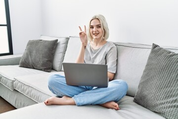 Poster - Young caucasian woman using laptop at home sitting on the sofa smiling with happy face winking at the camera doing victory sign. number two.