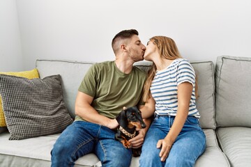 Wall Mural - Young hispanic couple kissing and sitting on the sofa with dog at home.