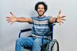 Handsome hispanic man sitting on wheelchair looking at the camera smiling with open arms for hug. cheerful expression embracing happiness.