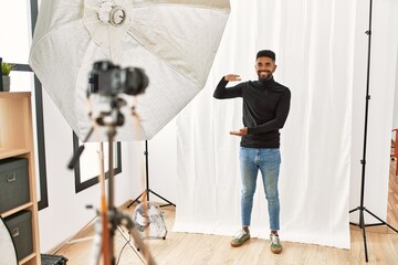 Wall Mural - Young hispanic man with beard posing as model at photography studio gesturing with hands showing big and large size sign, measure symbol. smiling looking at the camera. measuring concept.