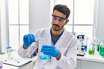 Wall Mural - Young hispanic man wearing scientist uniform pouring liquid on test tube at laboratory