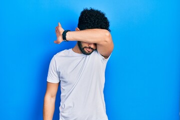 Young arab man with beard wearing casual white t shirt covering eyes with arm, looking serious and sad. sightless, hiding and rejection concept