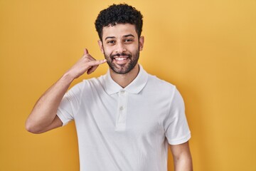 Sticker - Arab man standing over yellow background smiling doing phone gesture with hand and fingers like talking on the telephone. communicating concepts.