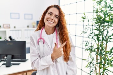 Sticker - Young redhead woman wearing doctor uniform and stethoscope at the clinic cheerful with a smile of face pointing with hand and finger up to the side with happy and natural expression on face