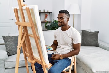 Wall Mural - Young african man painting on canvas at home looking to side, relax profile pose with natural face and confident smile.