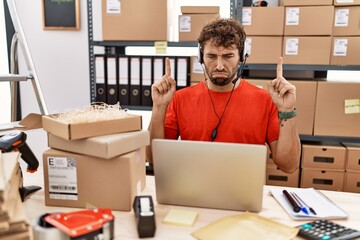 Poster - Young hispanic call center agent man working at warehouse pointing up looking sad and upset, indicating direction with fingers, unhappy and depressed.