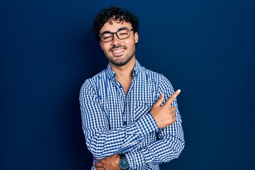 Canvas Print - Young hispanic man wearing casual clothes and glasses smiling cheerful pointing with hand and finger up to the side