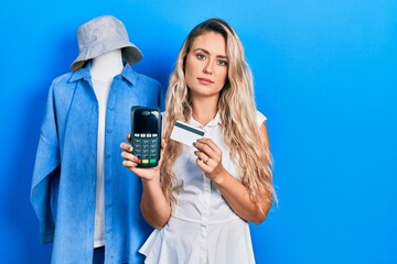 Poster - Beautiful young blonde shop assistance woman holding dataphone and credit card relaxed with serious expression on face. simple and natural looking at the camera.