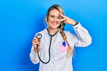 Sticker - Beautiful young blonde doctor woman holding stethoscope doing peace symbol with fingers over face, smiling cheerful showing victory