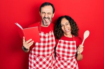 Wall Mural - Middle age couple of hispanic woman and man wearing professional apron reading cooking recipe book sticking tongue out happy with funny expression.