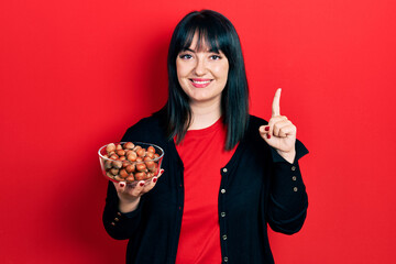 Sticker - Young hispanic woman holding bowl of chestnuts smiling with an idea or question pointing finger with happy face, number one