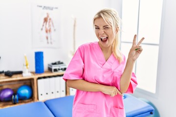 Poster - Young caucasian woman working at pain recovery clinic smiling with happy face winking at the camera doing victory sign. number two.