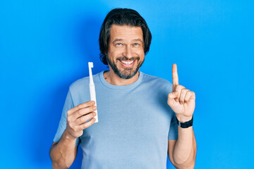 Canvas Print - Middle age caucasian man holding electric toothbrush smiling with an idea or question pointing finger with happy face, number one