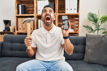 Young hispanic man with beard holding hammer and broken smartphone showing cracked screen angry and mad screaming frustrated and furious, shouting with anger looking up.